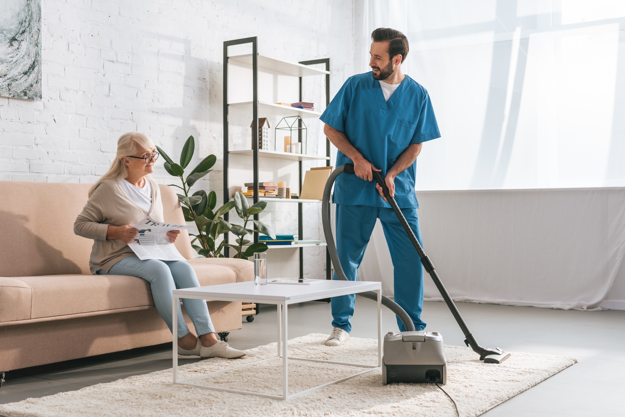 smiling social worker cleaning carpet with vacuum cleaner and looking at senior woman reading